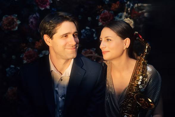 Clio Theodoridis, saxophone à la main, et Jonathan Nemtanu, du Duo Aster, posent l'un contre l'autre devant un fond floral sombre
