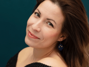 Portrait de Caroline Gélinas, souriante, la tête inclinée par-dessus l’épaule, devant un fond bleu-vert